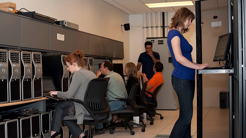 A server room aisle with cabinets of blinking equipment, cable trays overhead and a cold blue floor light.